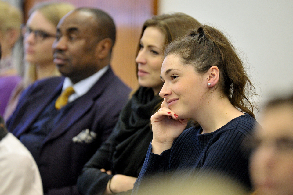 A young women, wearing a blue jumper, listening to a speech, sitting with other people.